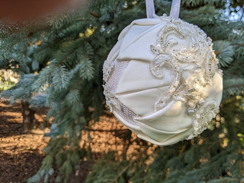 May include: A white fabric Christmas ornament with a beaded and lace design. The ornament is hanging from a tree branch.