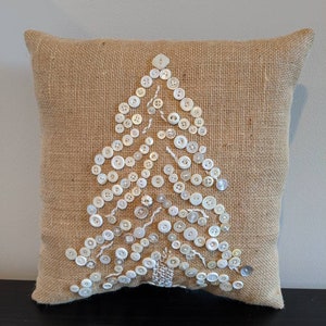 May include: A brown burlap cushion with a white button Christmas tree design. The buttons are sewn onto the cushion in the shape of a tree.