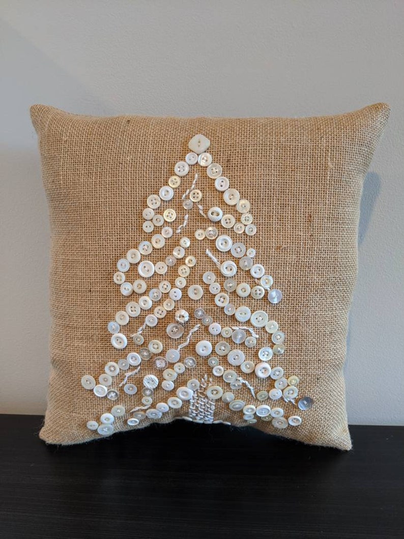 May include: A brown burlap cushion with a white button Christmas tree design. The buttons are sewn onto the cushion in the shape of a tree.
