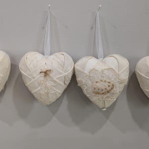 May include: Four white fabric heart-shaped ornaments with lace detailing. The hearts are hanging from white strings.