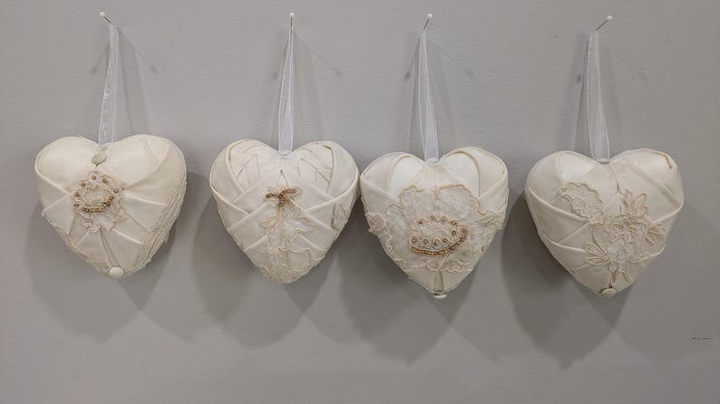 May include: Four white fabric heart-shaped ornaments with lace detailing. The hearts are hanging from white strings.