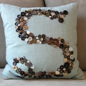 May include: A light blue throw pillow with a large letter "S" made of various sized buttons in brown, black, and white.
