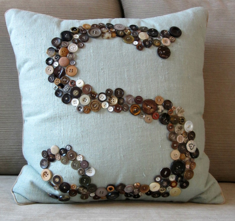 May include: A light blue throw pillow with a large letter "S" made of various sized buttons in brown, black, and white.