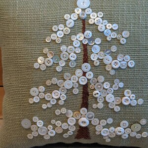 May include: A green burlap pillow with a white button tree design. The buttons are arranged in the shape of a tree with a brown embroidered trunk.