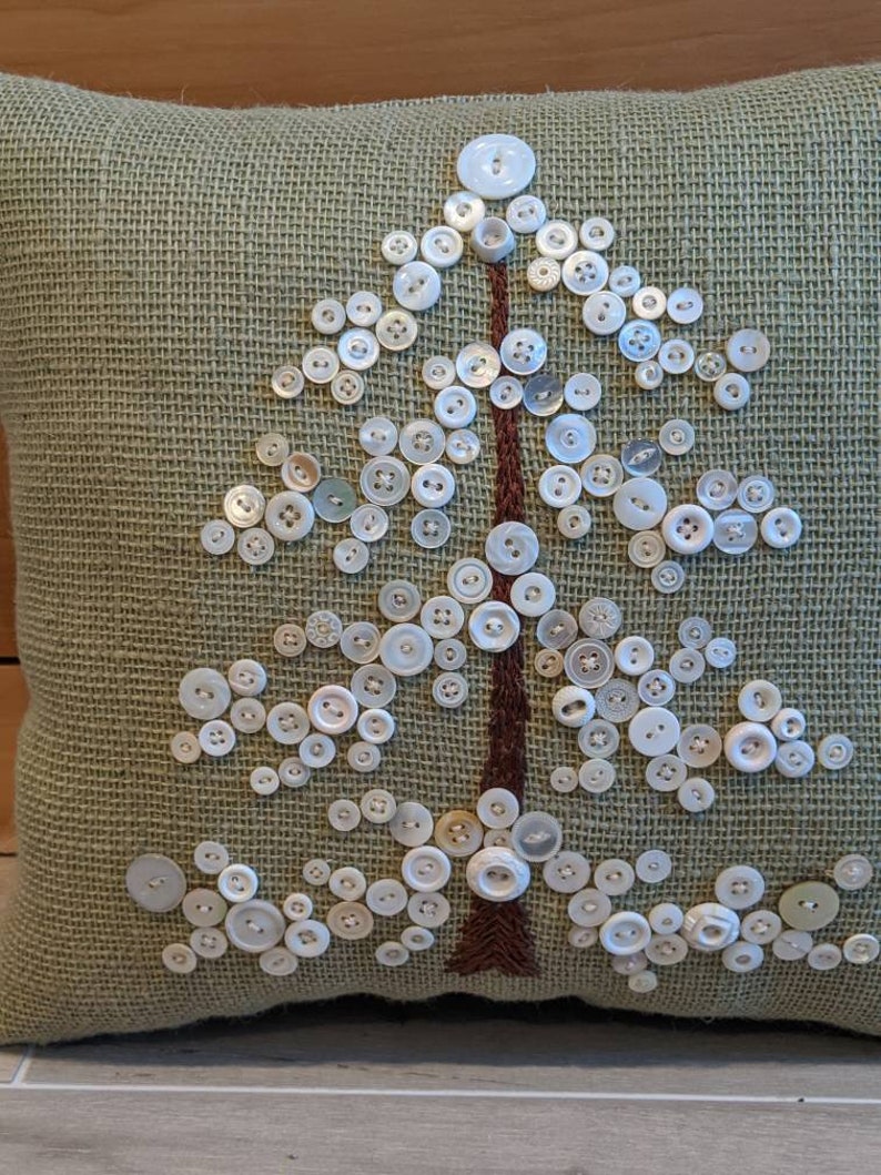 May include: A green burlap pillow with a white button tree design. The buttons are arranged in the shape of a tree with a brown embroidered trunk.