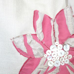 May include: Close-up of a white fabric with a large, decorative flower. The flower has pink and white striped petals and a center made of various sizes of white buttons. The flower is stitched onto the fabric.