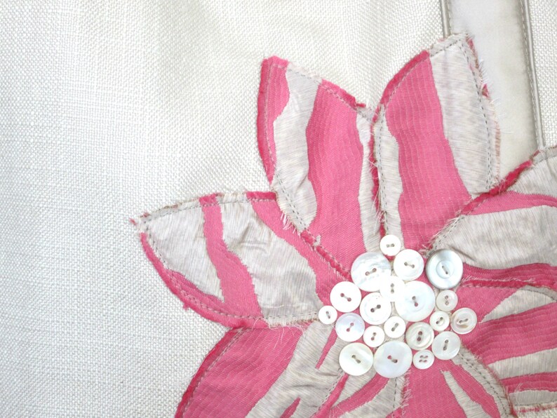 May include: Close-up of a white fabric with a large, decorative flower. The flower has pink and white striped petals and a center made of various sizes of white buttons. The flower is stitched onto the fabric.