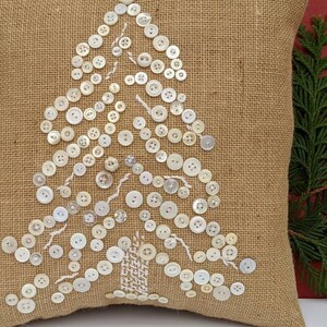 May include: A burlap cushion with a white button Christmas tree design. The cushion is a light brown colour and the buttons are white.