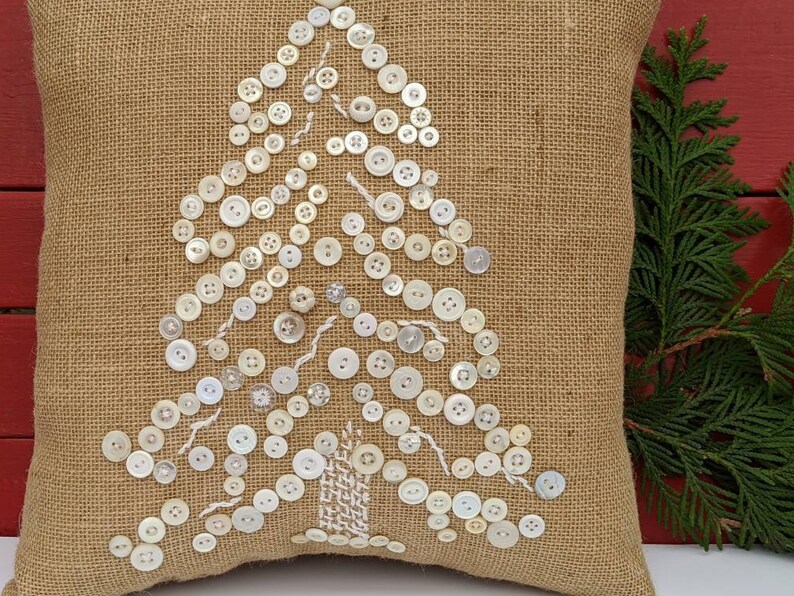May include: A burlap cushion with a white button Christmas tree design. The cushion is a light brown colour and the buttons are white.