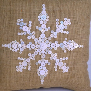 May include: A burlap pillow with a white snowflake design made of buttons.