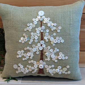 May include: A green burlap pillow with a white button tree design. The buttons are arranged in the shape of a tree with a brown stem.