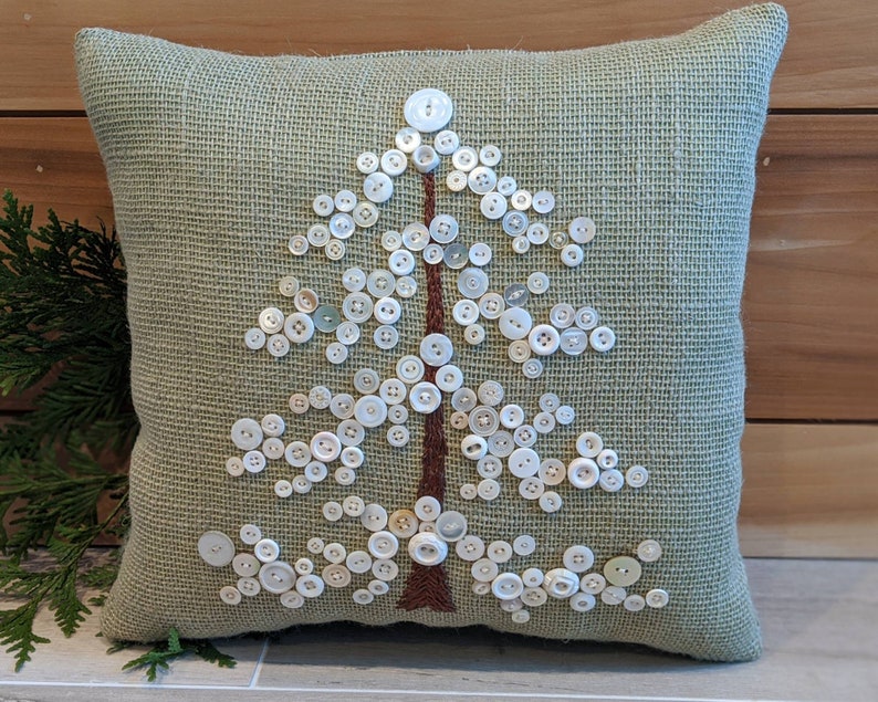 May include: A green burlap pillow with a white button tree design. The buttons are arranged in the shape of a tree with a brown stem.
