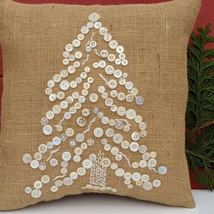 May include: A burlap cushion with a white button Christmas tree design. The cushion is a neutral beige colour and the buttons are white.