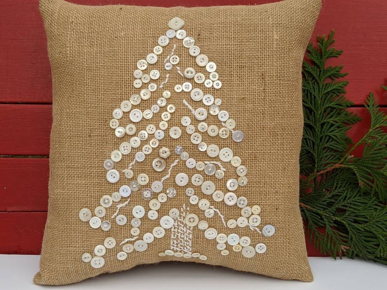 May include: A burlap cushion with a white button Christmas tree design. The cushion is a neutral beige colour and the buttons are white.