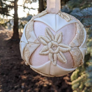 May include: A white and cream-colored fabric Christmas ornament with a floral design. The ornament is made of quilted fabric and has a delicate, embroidered flower in the center.