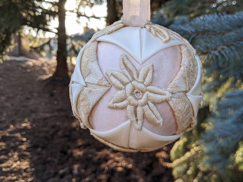 May include: A white and cream-colored fabric Christmas ornament with a floral design. The ornament is made of quilted fabric and has a delicate, embroidered flower in the center.