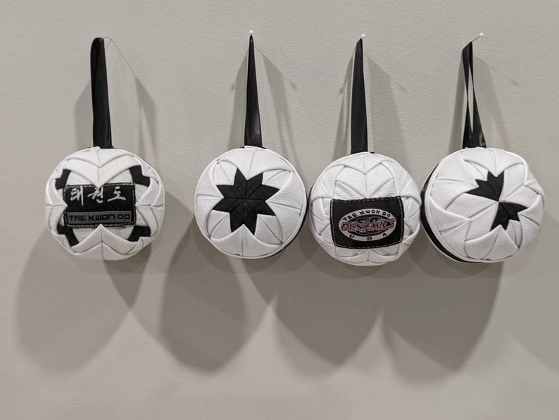 May include: Four white and black fabric ornaments with different designs hanging from black strings. The ornaments have a star, a Tae Kwon Do logo, and a Tae Kwon Do logo with the words "The Kwon Do of Punching" in a circle.