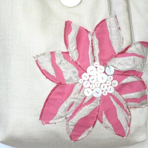 May include: A beige tote bag with a large, decorative flower applique. The flower features pink and white striped petals and a cluster of white buttons at the center. The bag has a button closure and a neutral-colored strap.