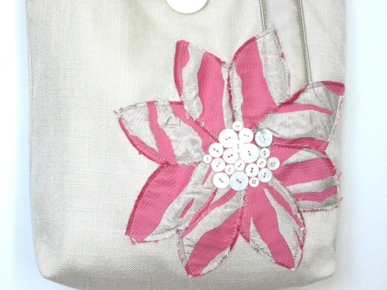 May include: A beige tote bag with a large, decorative flower applique. The flower features pink and white striped petals and a cluster of white buttons at the center. The bag has a button closure and a neutral-colored strap.
