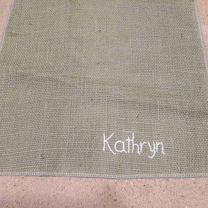 May include: A light green burlap placemat with the name "Kathryn" embroidered in white thread.