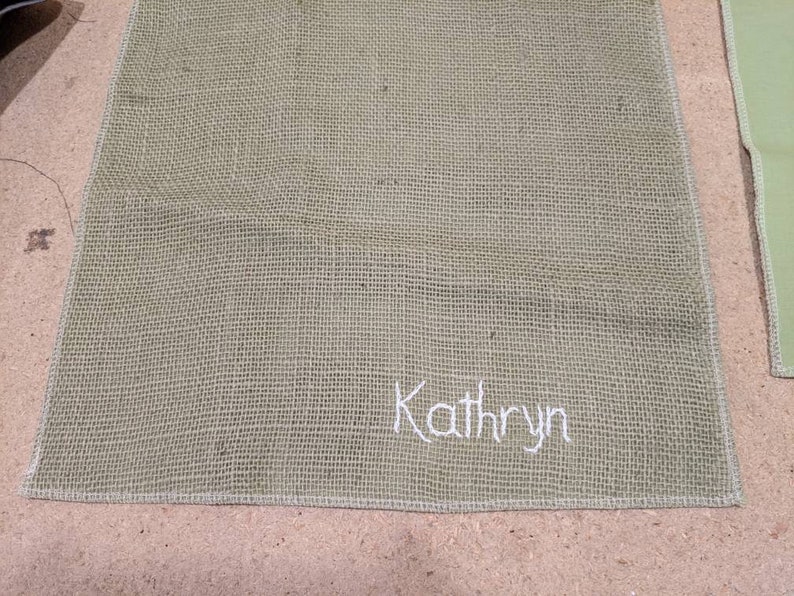 May include: A light green burlap placemat with the name "Kathryn" embroidered in white thread.