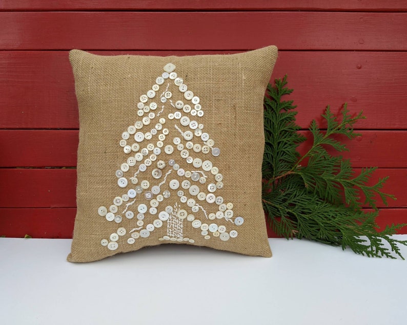 May include: A burlap cushion with a white button Christmas tree design. The cushion is on a white surface with a green branch in the background.