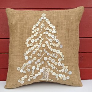 May include: A burlap cushion with a white button Christmas tree design. The cushion is a light brown colour and the buttons are white.