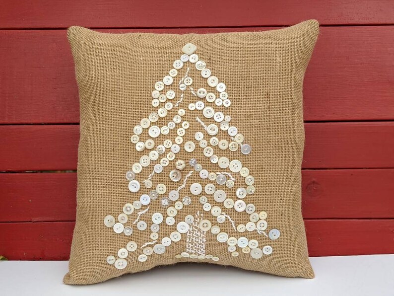 May include: A burlap cushion with a white button Christmas tree design. The cushion is a light brown colour and the buttons are white.