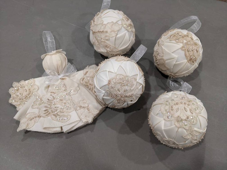 May include: Five white satin Christmas ornaments with lace and pearl embellishments. The ornaments are round and have a quilted design.