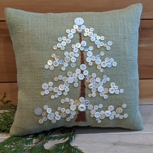 May include: A green throw pillow with a white button tree design. The pillow is made of a textured fabric and has a brown trim.