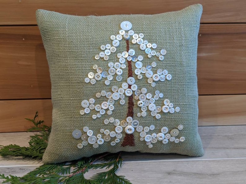 May include: A green throw pillow with a white button tree design. The pillow is made of a textured fabric and has a brown trim.