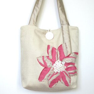 May include: Beige tote bag with a large pink and beige flower applique. The flower has pink petals with beige stripes and a cluster of white buttons at the center. The bag has beige handles and a white circular tag.