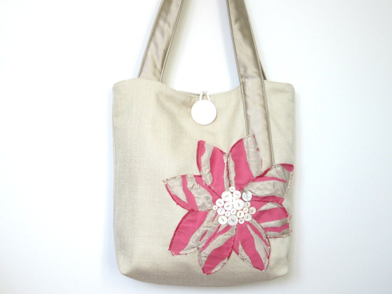 May include: Beige tote bag with a large pink and beige flower applique. The flower has pink petals with beige stripes and a cluster of white buttons at the center. The bag has beige handles and a white circular tag.
