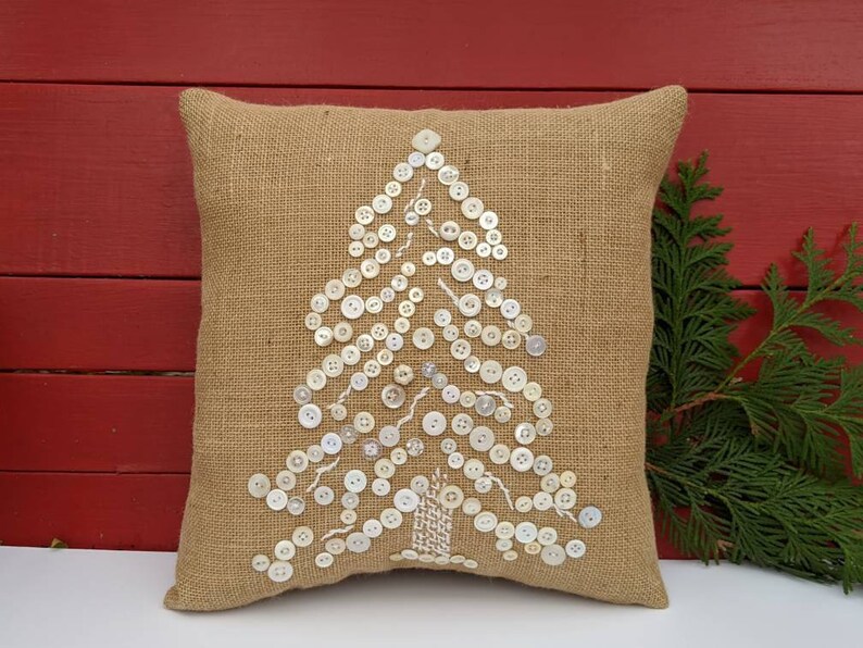 May include: A brown burlap cushion with a white button Christmas tree design. The cushion is on a red wooden background.
