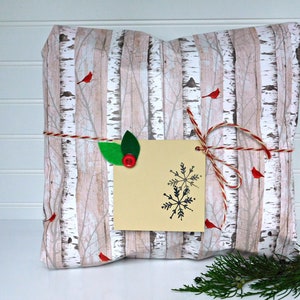 May include: A gift wrapped in birch tree patterned paper with red robins and a snowflake gift tag. The gift is tied with twine and decorated with green leaves.