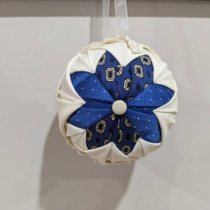 May include: A white fabric ornament with a blue and gold patterned flower design. The ornament has a loop for hanging.