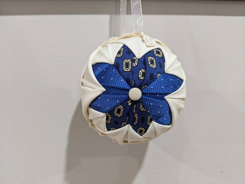 May include: A white fabric ornament with a blue and gold patterned flower design. The ornament has a loop for hanging.