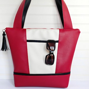 May include: A red and white tote bag with black trim and a black tassel. The bag has a zippered pocket on the front and a pair of sunglasses are hanging from the zipper.