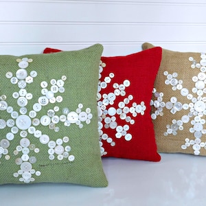 Puede incluir: Tres almohadas decorativas con diseños de copos de nieve hechos con botones blancos sobre telas de diferentes colores: verde, rojo y marrón.
