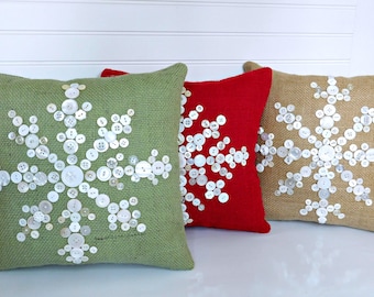 May include: Three decorative throw pillows with snowflake designs made from white buttons on different colored fabrics: green, red, and brown.