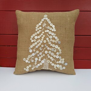 May include: A brown burlap cushion with a white button Christmas tree design. The buttons are sewn onto the cushion in the shape of a tree.