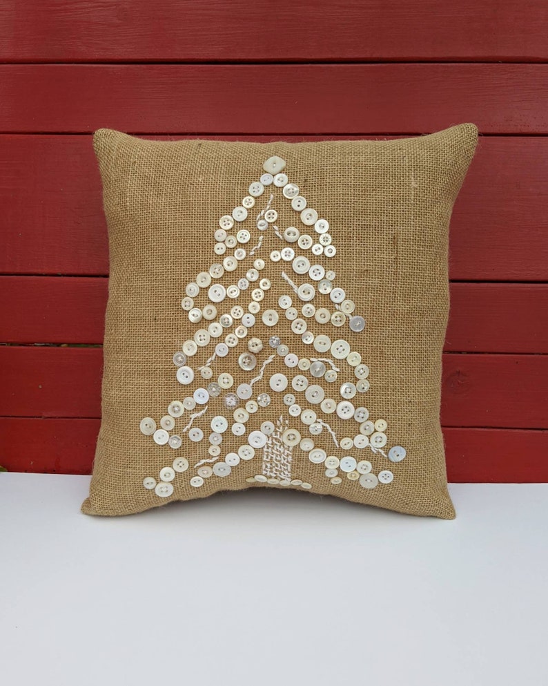 May include: A brown burlap cushion with a white button Christmas tree design. The buttons are sewn onto the cushion in the shape of a tree.