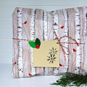 May include: A gift wrapped in birch tree patterned paper with red cardinals and a snowflake gift tag. The gift is tied with twine and decorated with green leaves.