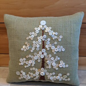 May include: A green burlap pillow with a white button tree design. The buttons are arranged in the shape of a tree with a brown embroidered trunk.