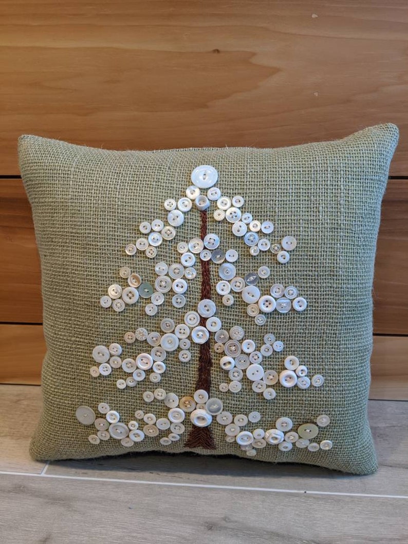 May include: A green burlap pillow with a white button tree design. The buttons are arranged in the shape of a tree with a brown embroidered trunk.