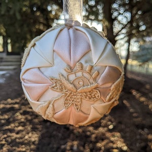 May include: A handmade, quilted Christmas ornament in shades of pink and white with gold embroidery. The ornament is shaped like a sphere and has a satin finish.