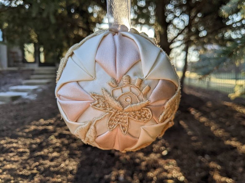 May include: A handmade, quilted Christmas ornament in shades of pink and white with gold embroidery. The ornament is shaped like a sphere and has a satin finish.