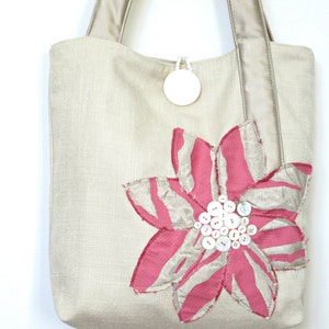May include: A beige tote bag with a large pink and silver flower applique. The flower is embellished with white buttons at the center. The bag has beige handles and a small white circular tag attached to the top.
