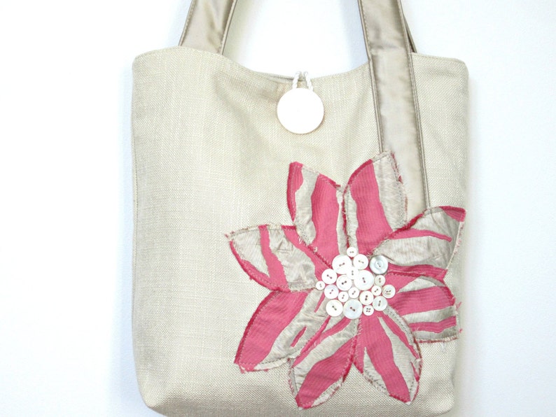 May include: A beige tote bag with a large pink and silver flower applique. The flower is embellished with white buttons at the center. The bag has beige handles and a small white circular tag attached to the top.