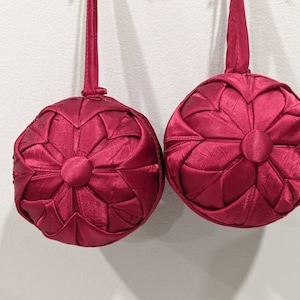 May include: Two burgundy fabric Christmas ornaments with a starburst design. The ornaments are hanging from a white wall.